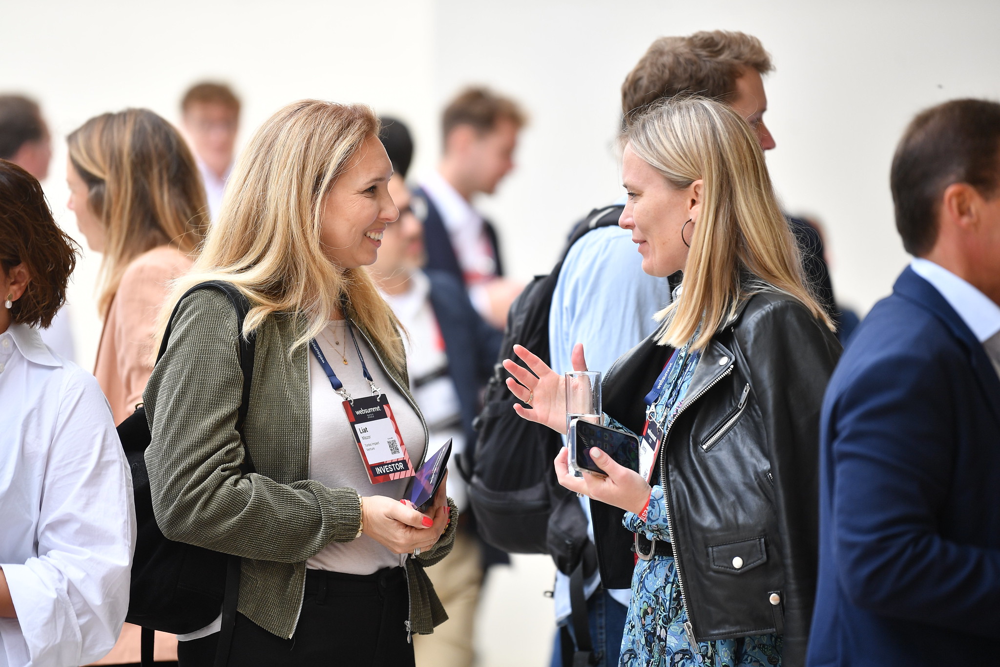 Two smiling people face each other in a crowd. Both are holding mobile devices in their hands. The person on the right has raised one hand as if emphasising a point. They appear to be in conversation.