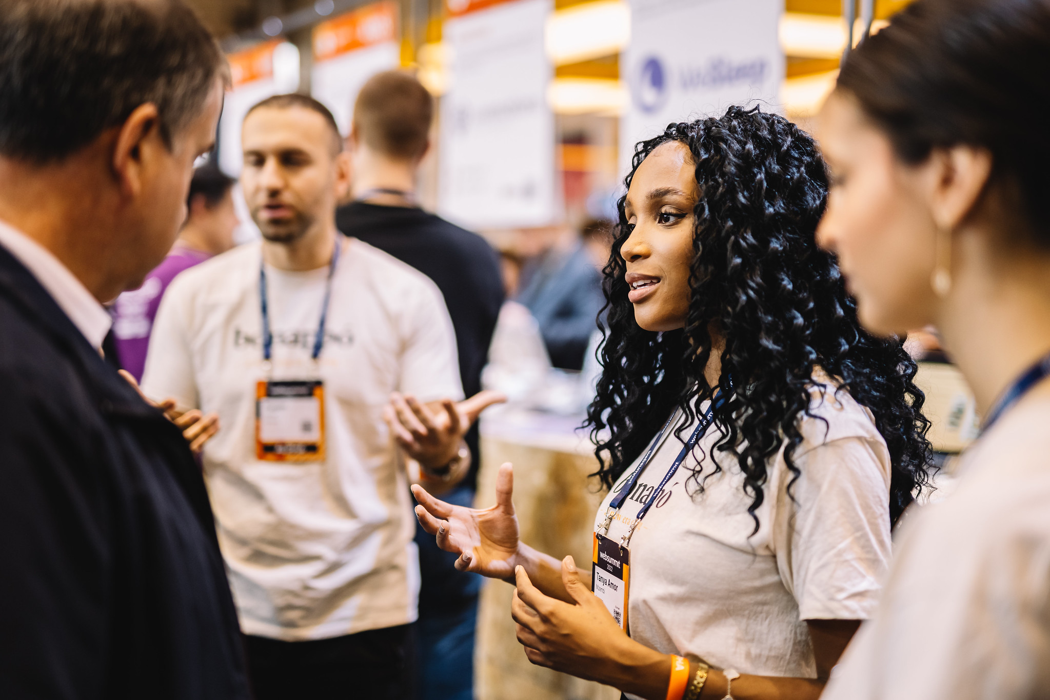 A person appears to be addressing a small crowd of people. The person is gesturing with both hands as if emphasising a point. Visible in the background are people and exhibition stands.